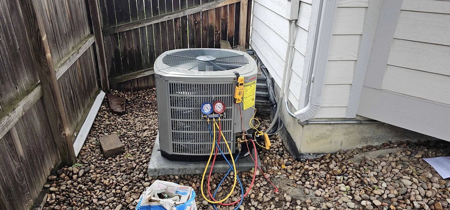 Outdoor condenser on a pad in a fenced gravel side yard with red and blue refrigerant gauges attached