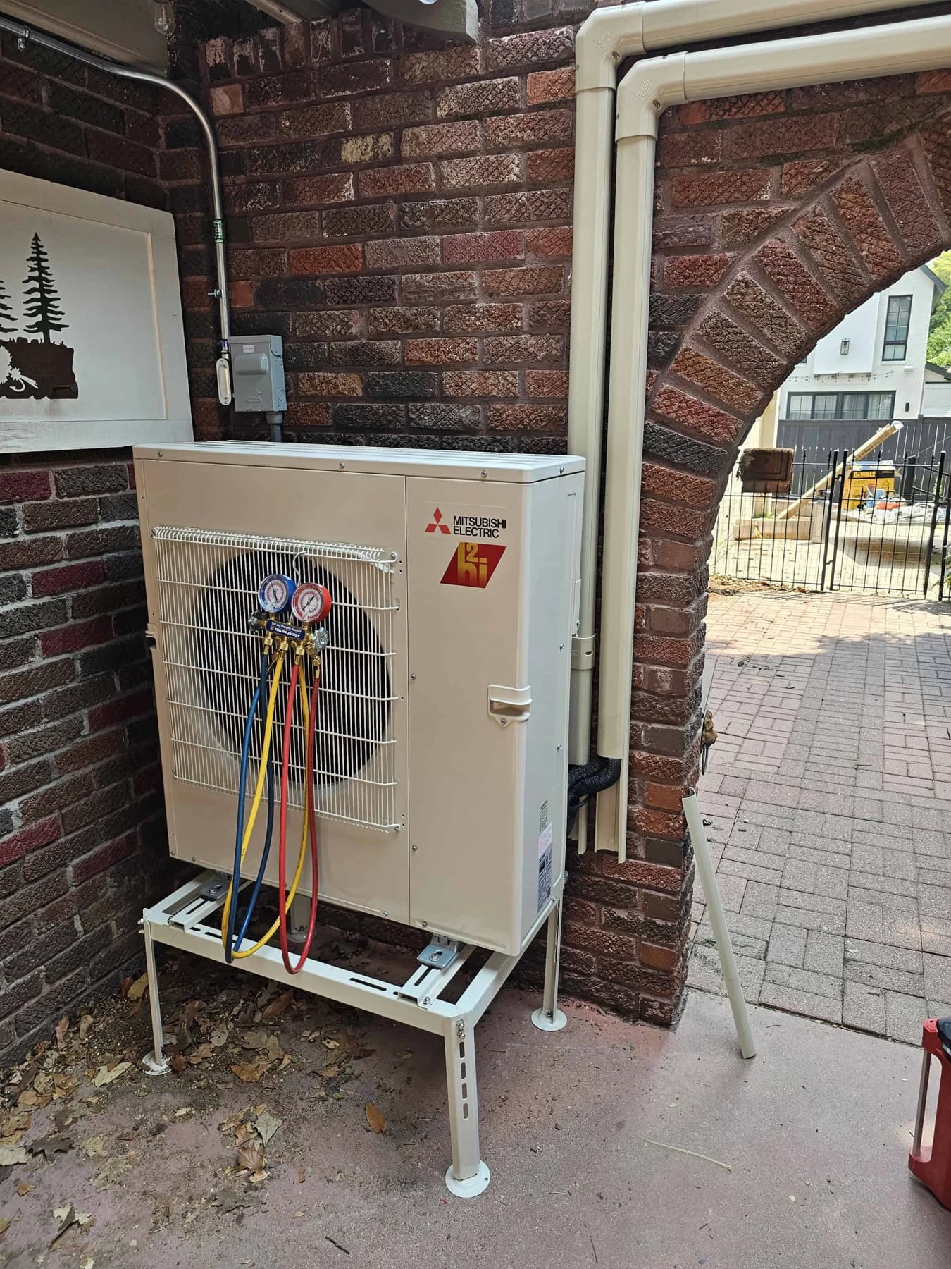 Outdoor heat pump with gauges, disconnect, and covered lines beside a brick arch