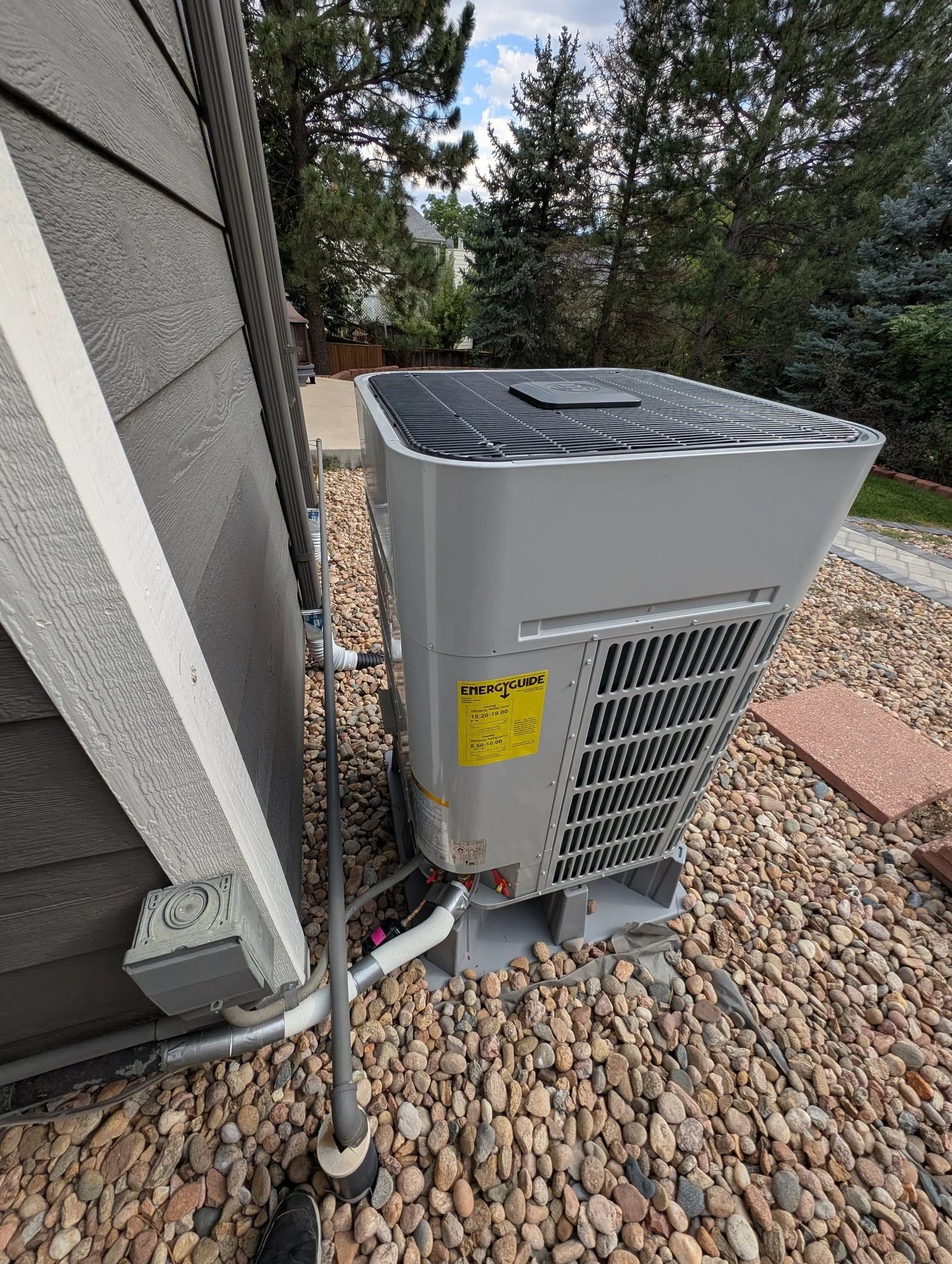 A Bosch heat pump on a composite pad in a river-rock bed, with pine trees behind