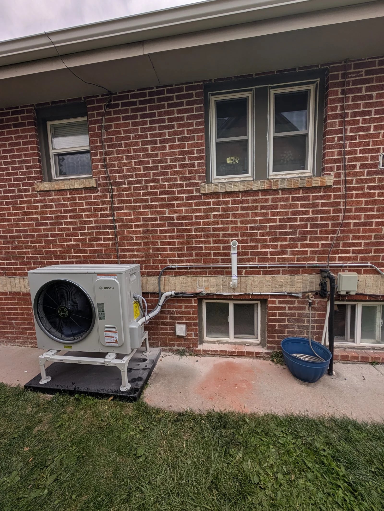A Bosch heat pump on a stand on the lawn beside a red brick ranch house