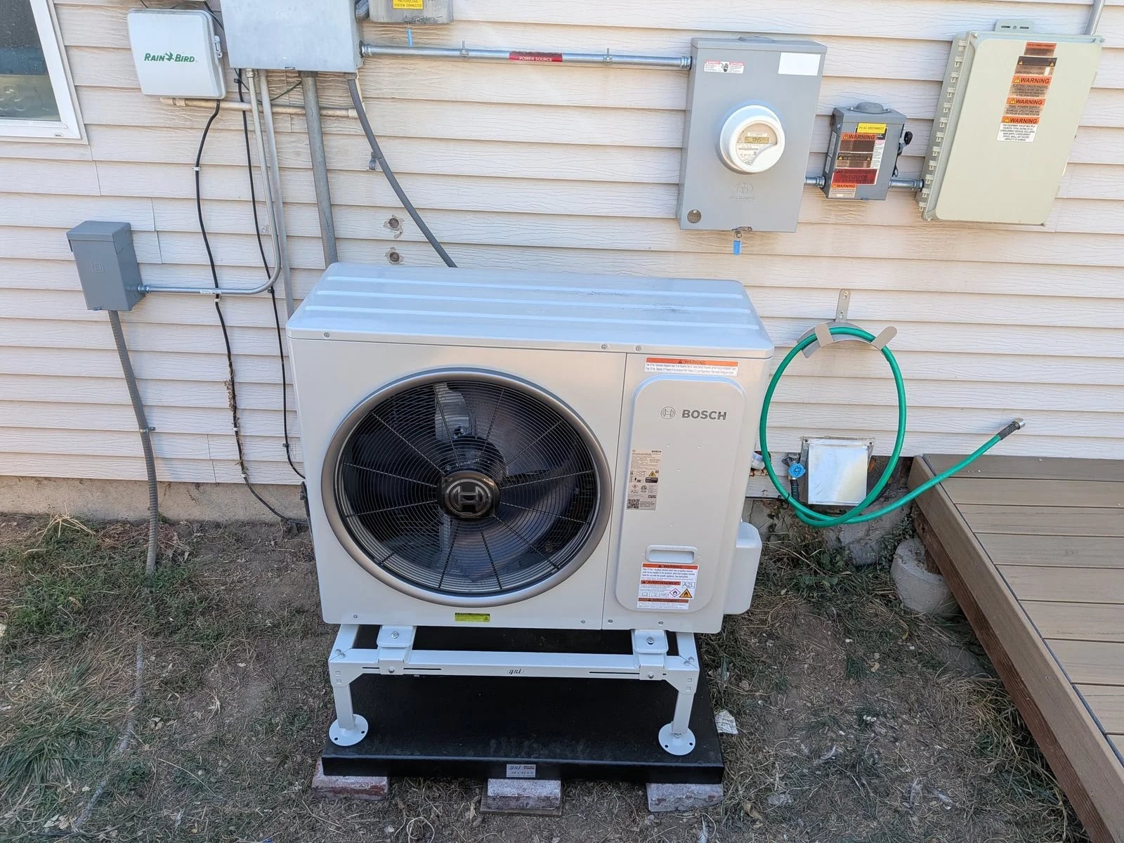 A Bosch heat pump on a raised stand below the electric meter, panel and disconnects