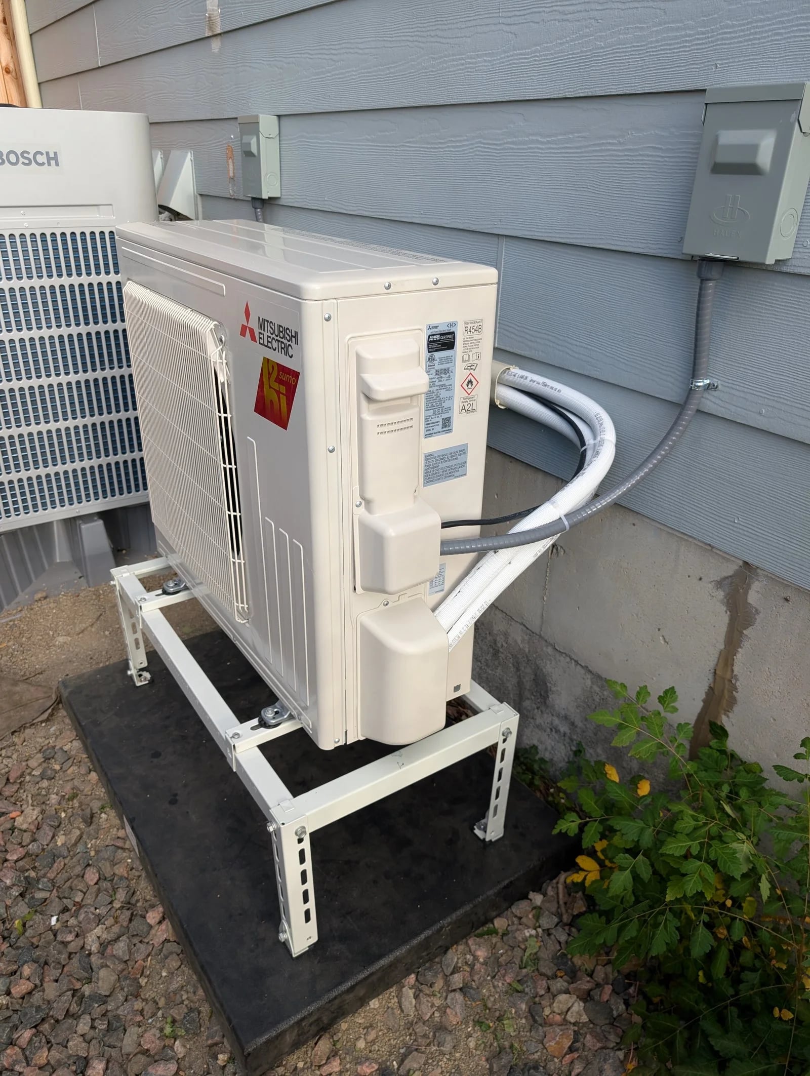 A ductless outdoor unit on a stand, its line set running into the wall beside a Bosch heat pump