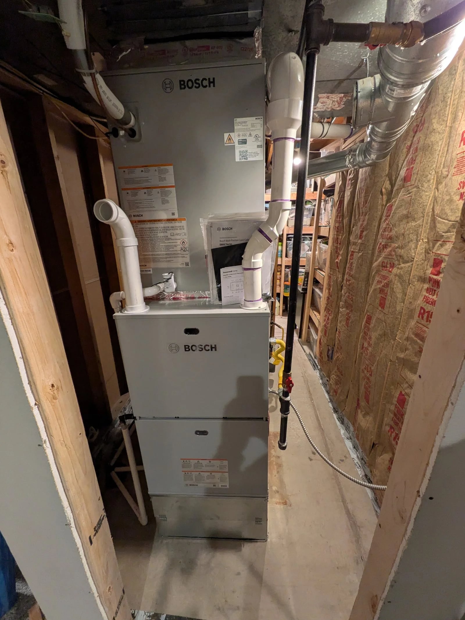 A Bosch gas furnace and coil with white PVC intake and exhaust pipes in a narrow basement space
