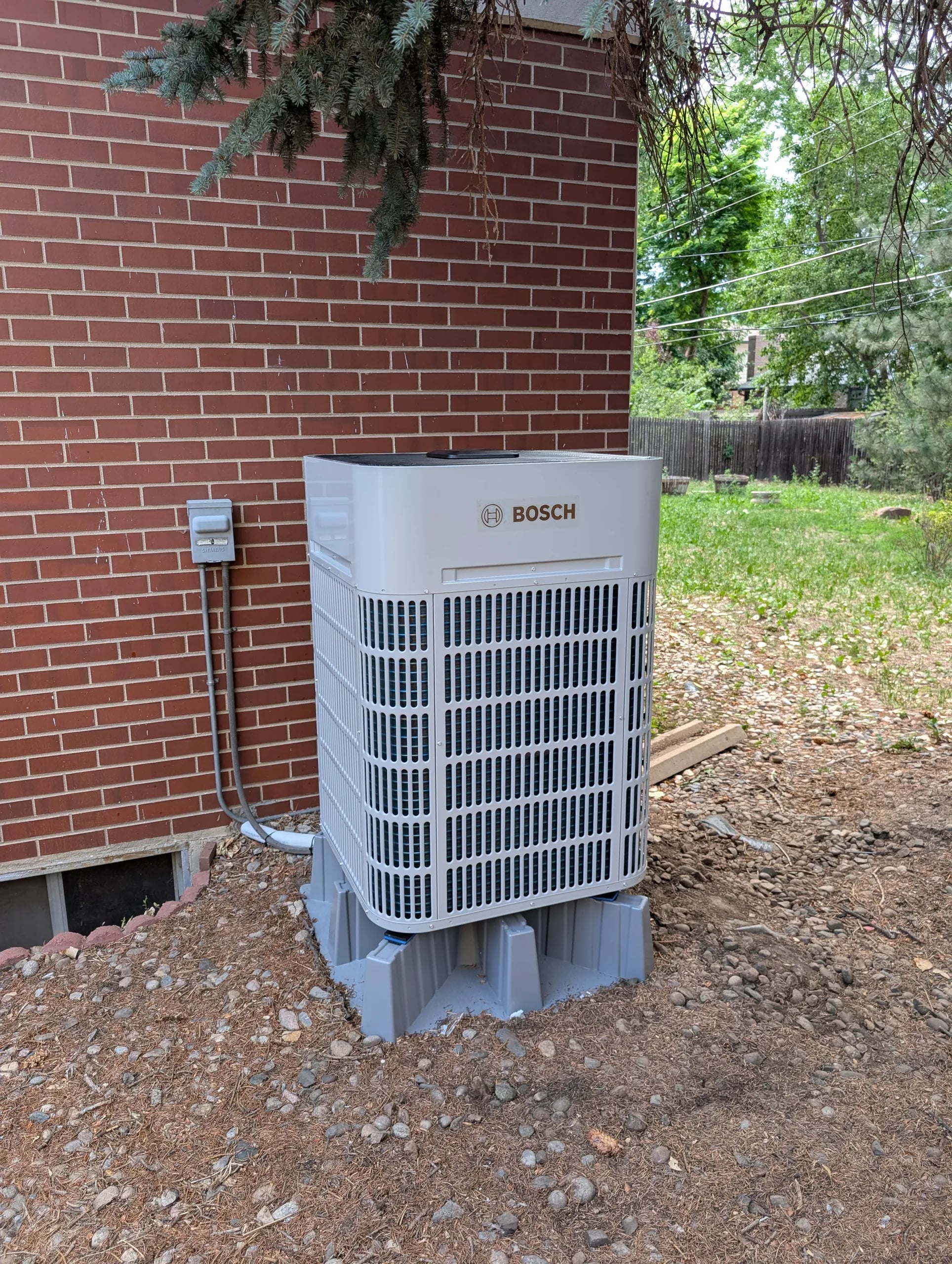 Bosch condenser on risers on bare soil beside a red brick wall, a tree behind it