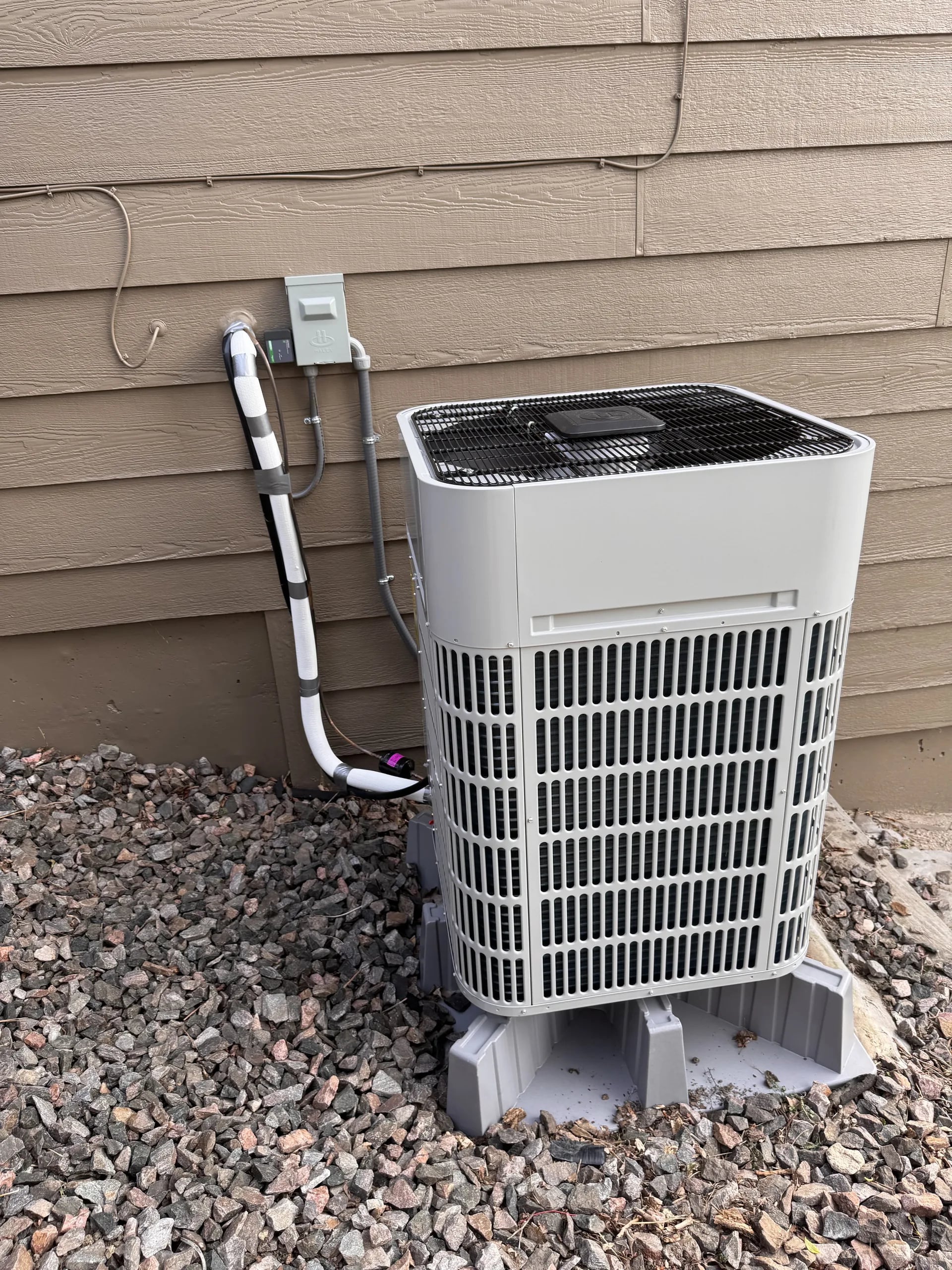 Gray condenser on risers in red gravel beside brown siding, its line-set cover rising to a disconnect