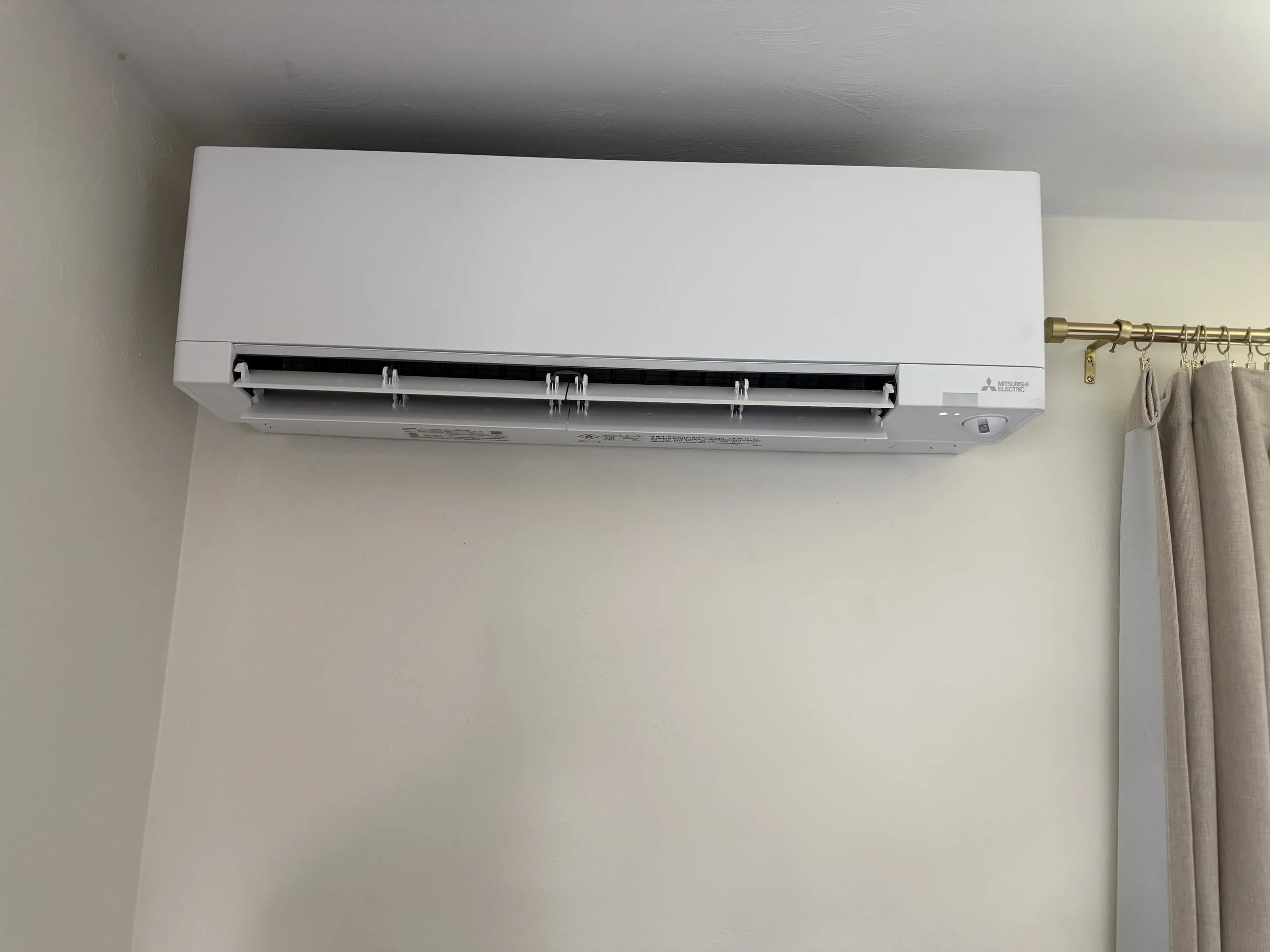 White Mitsubishi wall-mounted head high on a white wall beside a curtain rod